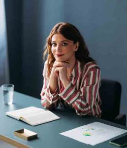 Professional female entrepreneur in a striped shirt at work, focused with notebook and papers on her desk. Completing an AI readiness assessment provided by an MSP as part of their AI services.